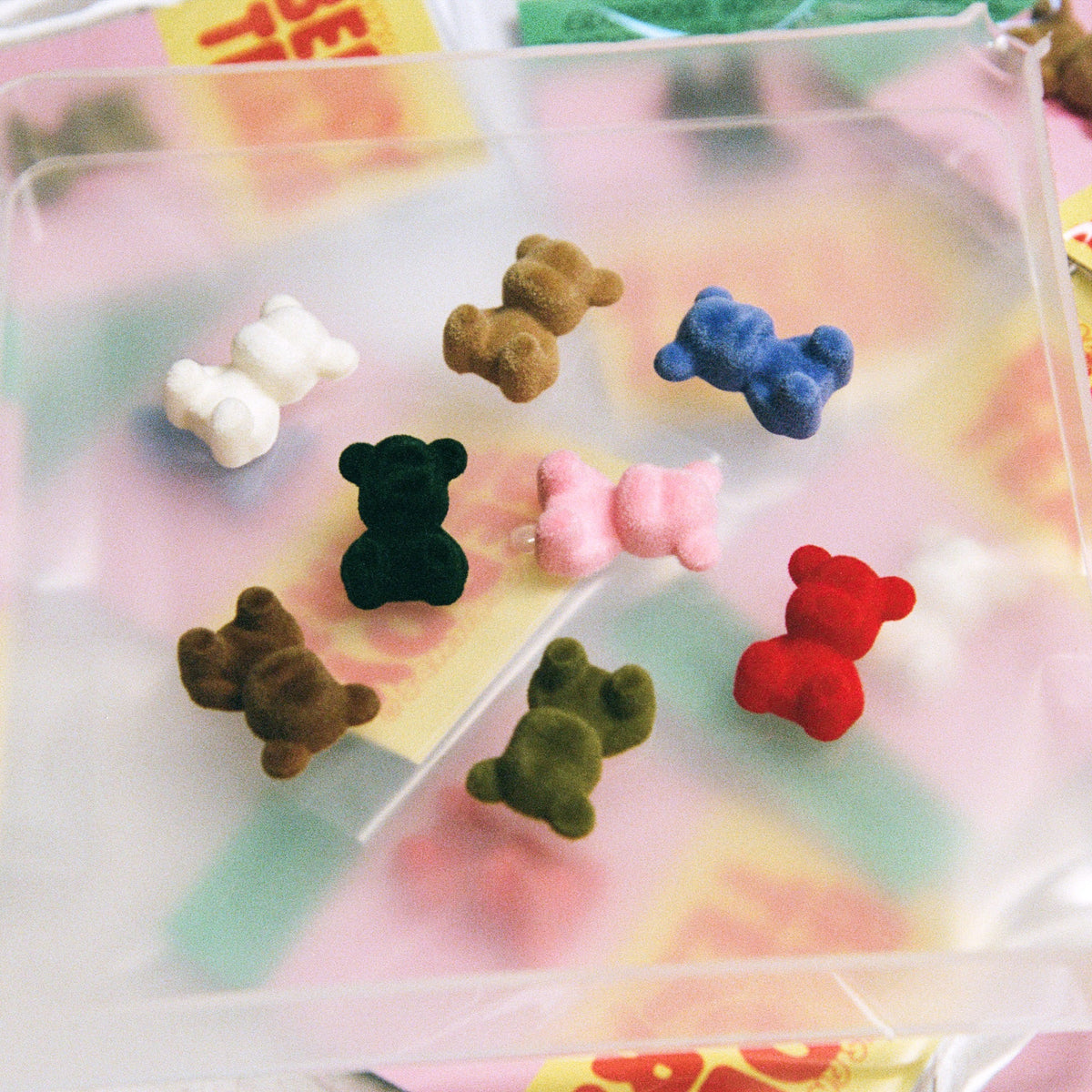 A clear plastic tray holding seven small teddy bear-shaped items in assorted colors, including pink, brown, blue, black, light pink, red, and green, arranged on top of colorful candy wrappers.