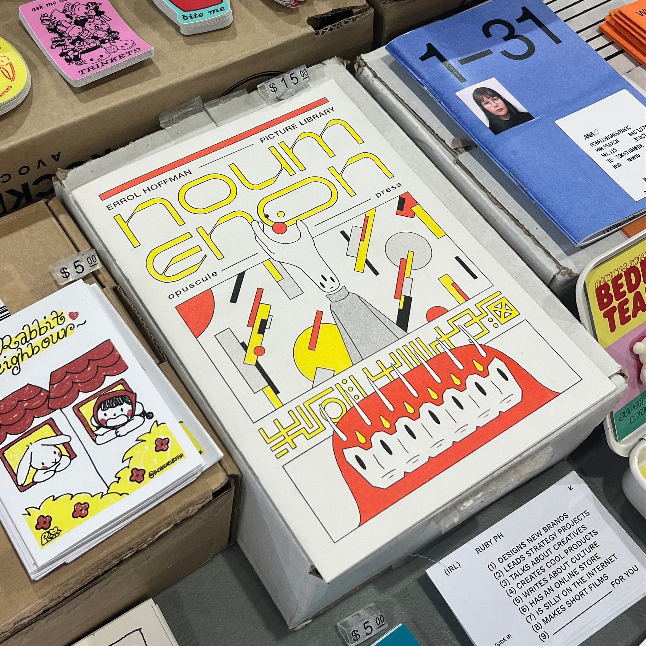 A tabletop display of various stationery items, including notebooks, a planner, stickers, and cards, with one prominent white notebook featuring the text 'noumea' and colorful illustrations.