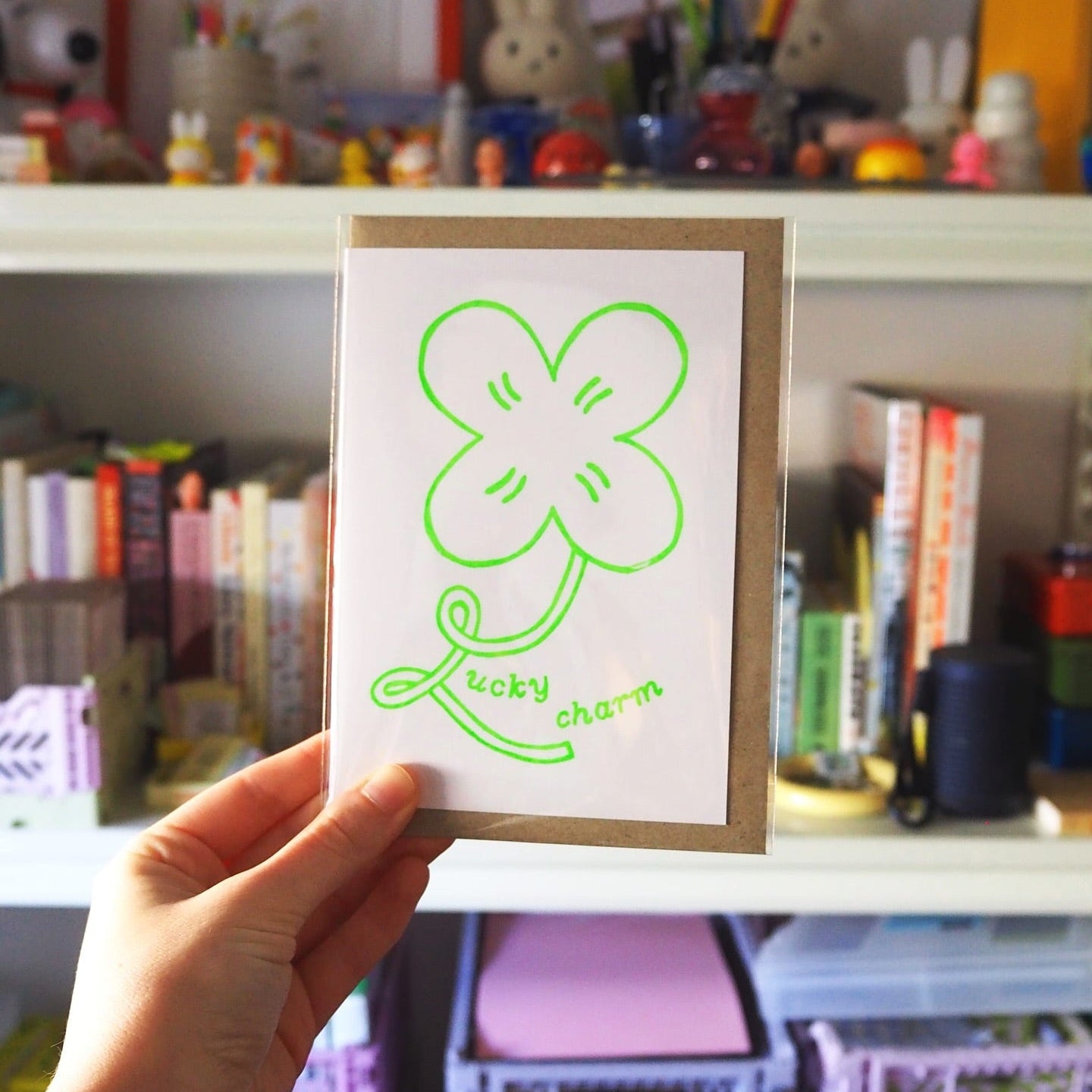 A hand holds a small greeting card in front of a blurred bookshelf. The card has a white background with a bright green outline drawing of a four-leaf clover and the words 'Lucky charm' in green script.