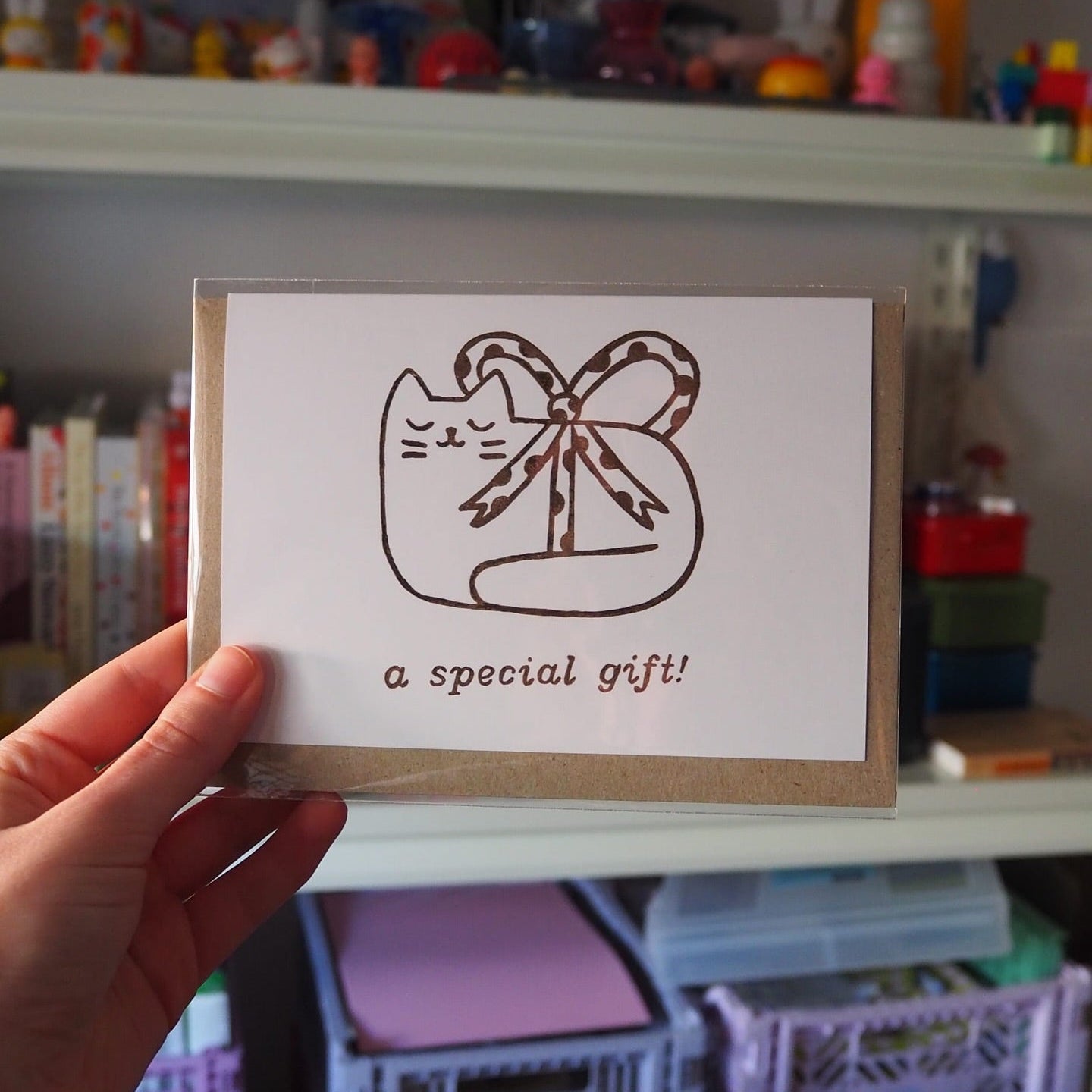 A hand holds a square greeting card in front of a blurred shelf background. The card has a white front with a brown line drawing of a cat-shaped gift box tied with a bow and the text 'a special gift!' below it.