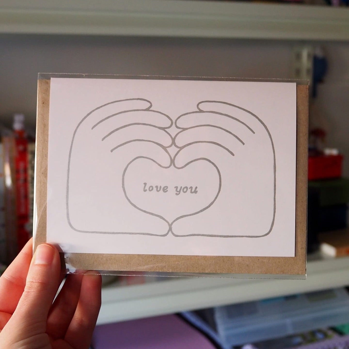 A hand holds a small greeting card with a simple line drawing of two hands forming a heart shape and the words 'love you' in the center. The card is wrapped in clear plastic and placed over a kraft envelope, with shelves of colorful items blurred in the background.